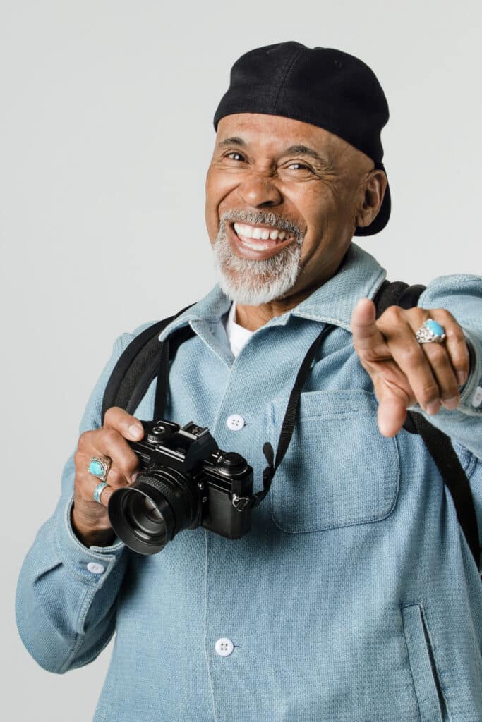 A smiling man in a blue jacket points playfully while holding a camera.