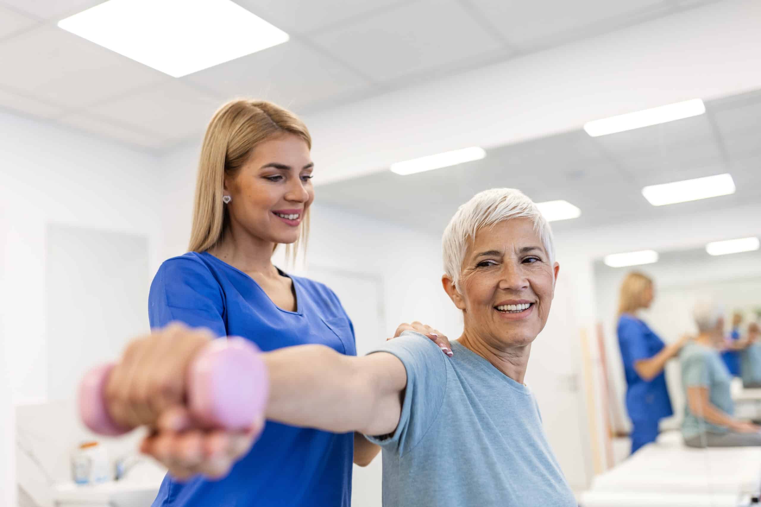 A therapist helps a smiling older woman lift a pink dumbbell during exercise.