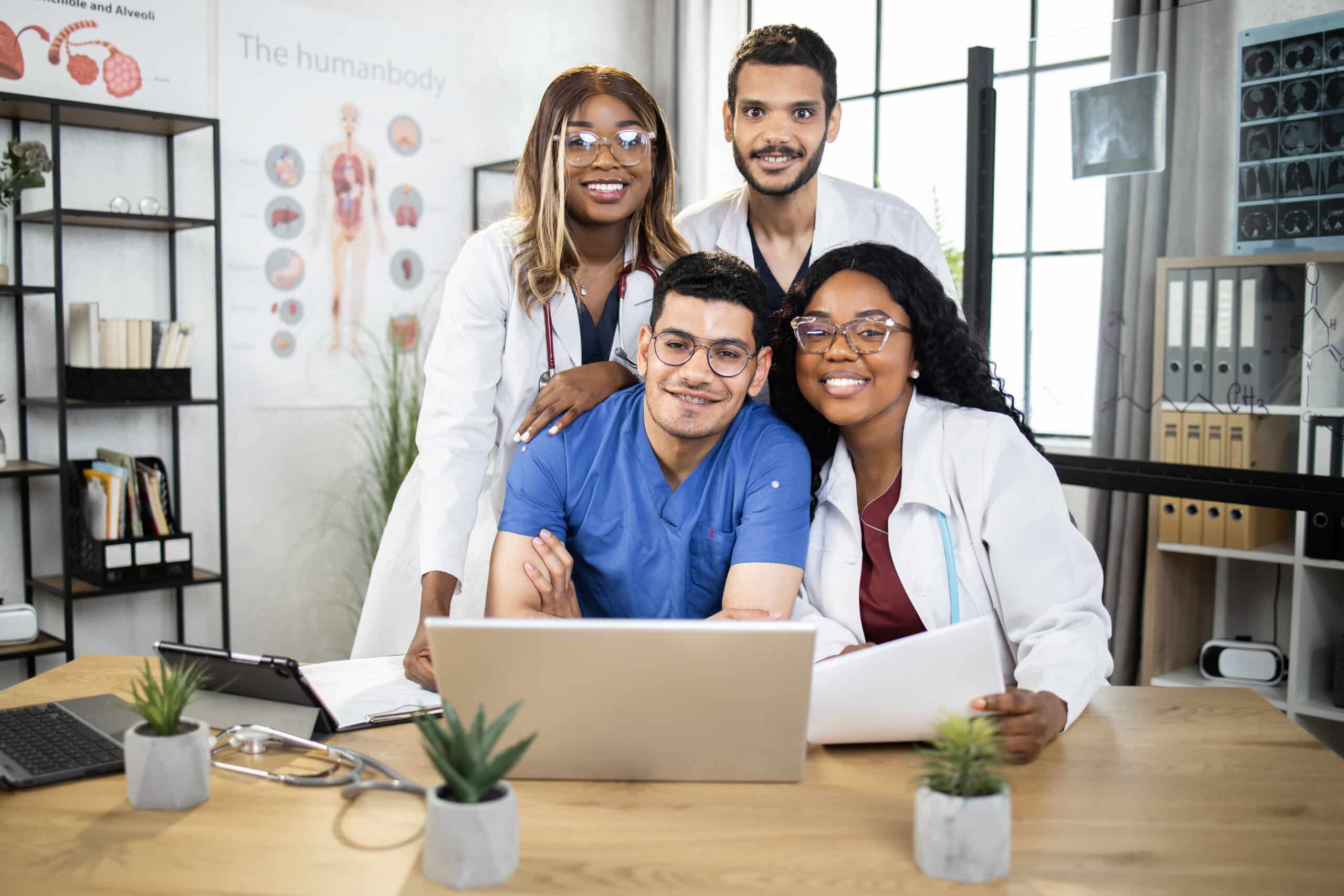 Group of four international doctors in lab coat and scrubs sitting together at table and using laptop and tablet. Medical conference of male and female specialists. Sharing with knowledges.