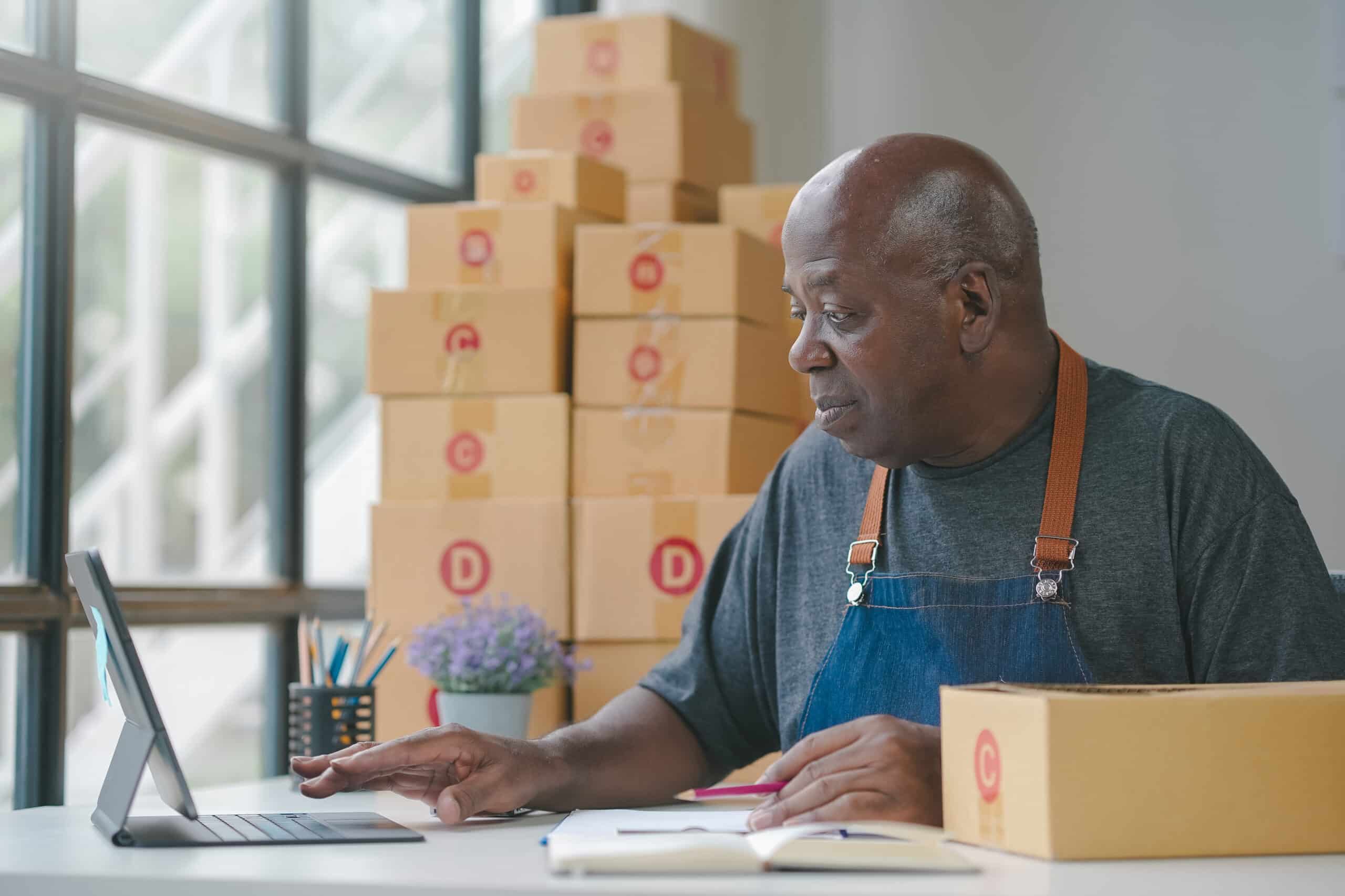 Senior man in workshop working on a laptop with boxes stacked in the background, focused and diligent in his task.