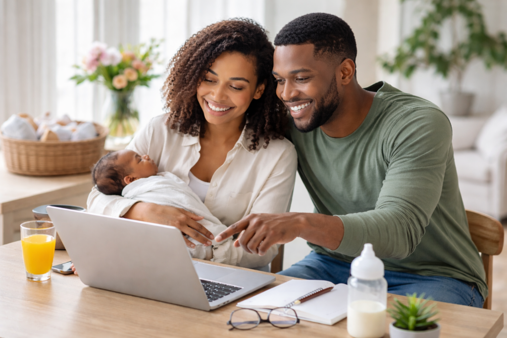 Two adults smiling at a laptop while one holds a baby wrapped in a white blanket.