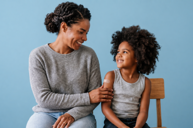 An adult and a child sit side by side against a light blue background. The adult, wearing a gray sweater, gently touches the child’s arm, which has a small adhesive bandage, suggesting the child recently received a vaccination. Both are smiling warmly, conveying comfort and care