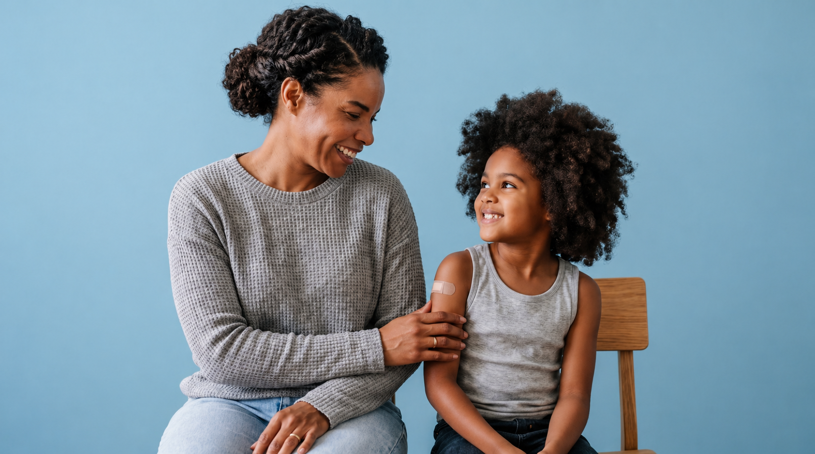An adult and a child sit side by side against a light blue background. The adult, wearing a gray sweater, gently touches the child’s arm, which has a small adhesive bandage, suggesting the child recently received a vaccination. Both are smiling warmly, conveying comfort and care